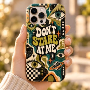 May include: A phone case featuring a psychedelic design with the text "DON'T STARE AT ME" in a wavy font. The artwork includes multiple eyes, stars, a planet with rings, and a black and white checkerboard pattern in shades of green, orange, and black.