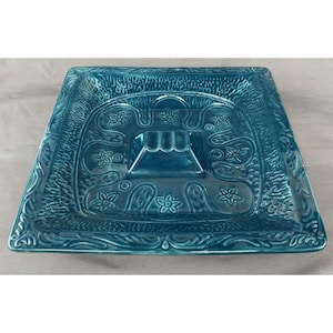 May include: A square teal ceramic dish featuring a central raised square pedestal with three indentations. The surface is decorated with embossed floral patterns and swirling lines, framed by a textured border along the outer edges.