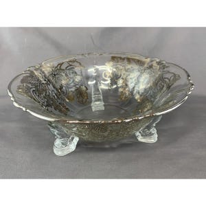 May include: A clear glass bowl featuring a scalloped rim and three decorative feet. The exterior surface is adorned with a metallic gold leaf pattern depicting intricate grapevines, leaves, and swirling scrollwork designs.