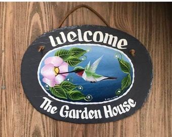 May include: A black slate oval sign with a white painted welcome sign, a painted hummingbird with green and red feathers, a pink flower, and green leaves. The sign also says "The Garden House" in white paint.