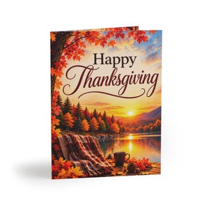 May include: A greeting card featuring a sunset over a lake with autumn trees, a plaid blanket draped over a wooden chair, and a mug. The text Happy Thanksgiving is printed in dark brown script and serif font at the top of the card.