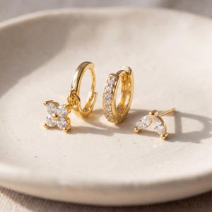 Puede incluir: Un conjunto de tres pendientes dorados que incluye un aro con un dije de flor de cuatro pétalos, un aro pequeño con piedras transparentes incrustadas y un pendiente de botón con tres piedras transparentes en forma de abanico.