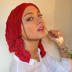 May include: A woman wearing a red ruffled head scarf. The scarf is made of a soft, flowing fabric and has a gathered, ruffled design. The woman is looking to the side and has a neutral expression on her face.