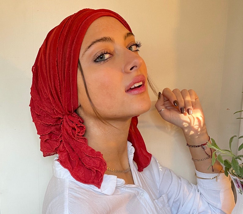 May include: A woman wearing a red ruffled head scarf. The scarf is made of a soft, flowing fabric and has a gathered, ruffled design. The woman is looking to the side and has a neutral expression on her face.