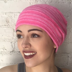 May include: A pink striped beanie hat. The hat is made of a soft, textured material and features alternating shades of pink. The beanie is worn by a person with dark hair and a gray tank top. The background is a white brick wall.
