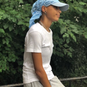 May include: A person wearing a light blue bandana tied around their head as a headband, a light blue baseball cap, a white t-shirt, and gray sweatpants. The person is standing in front of a green leafy background.