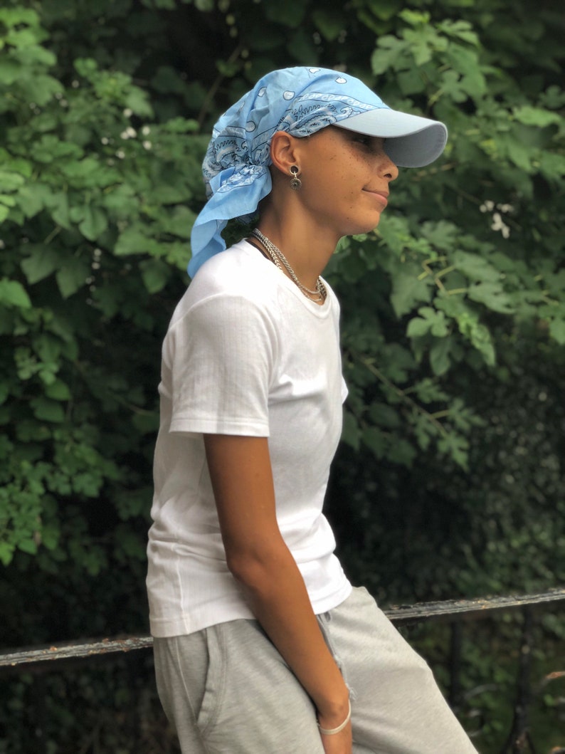 May include: A person wearing a light blue bandana tied around their head as a headband, a light blue baseball cap, a white t-shirt, and gray sweatpants. The person is standing in front of a green leafy background.