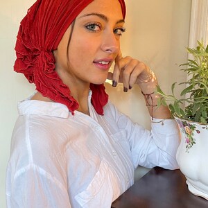 May include: A woman wearing a red ruffled head scarf and a white button-down shirt. She is leaning on a wooden table.