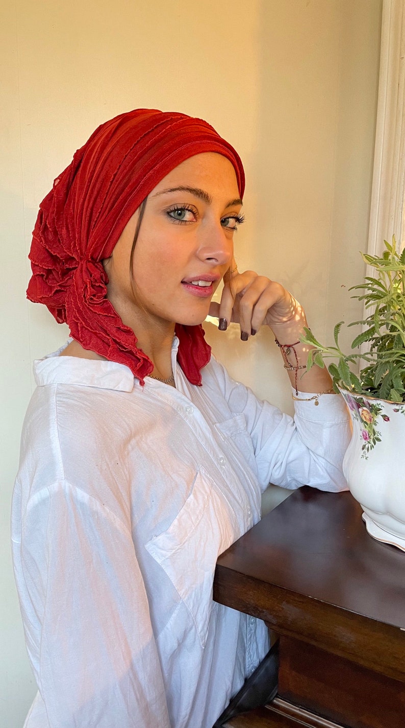 May include: A woman wearing a red ruffled head scarf and a white button-down shirt. She is leaning on a wooden table.