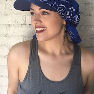 May include: A woman wearing a blue bandana with a white pattern and a blue baseball cap with a visor. She is wearing a gray tank top with a Nike logo.