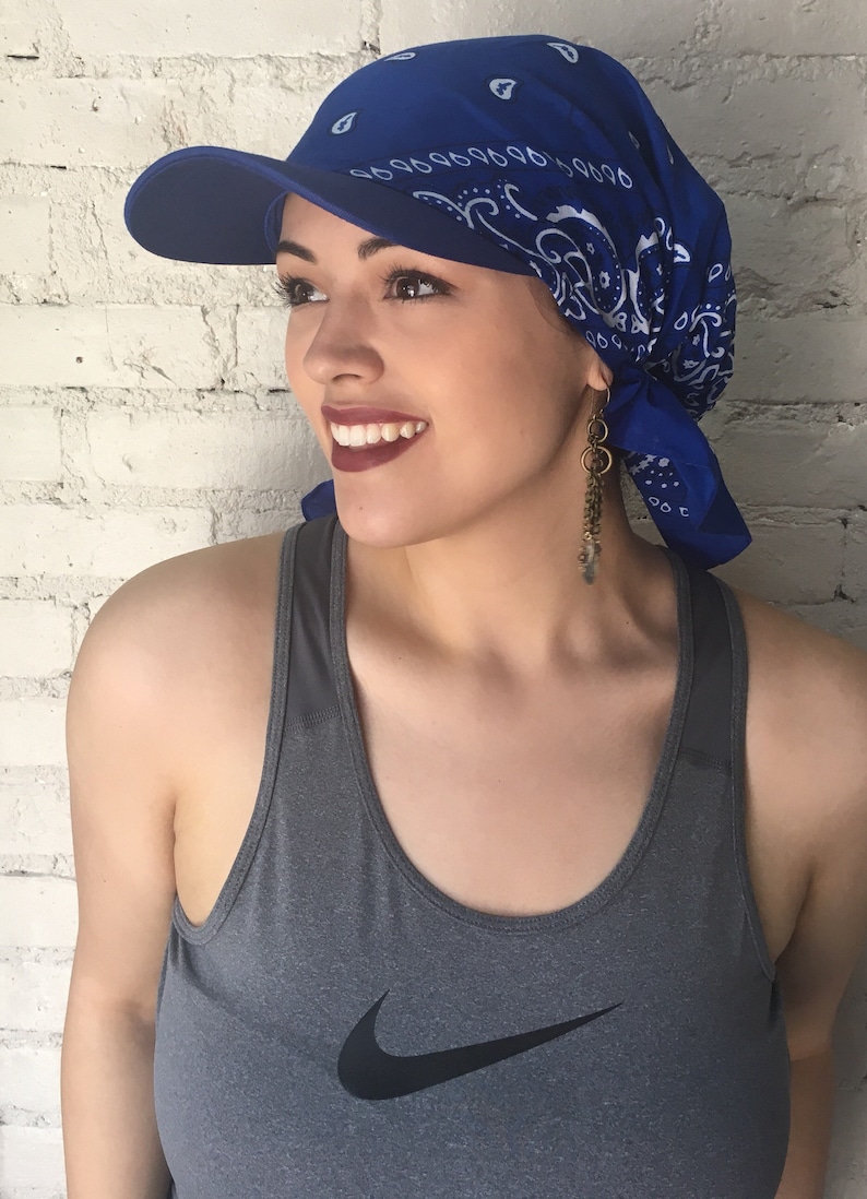 May include: A woman wearing a blue bandana with a white pattern and a blue baseball cap with a visor. She is wearing a gray tank top with a Nike logo.