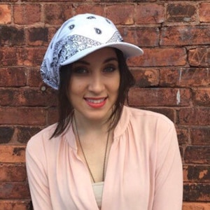 May include: A white bandana style baseball cap with a black paisley pattern. The cap is worn by a person with long brown hair and they are wearing a light pink shirt.