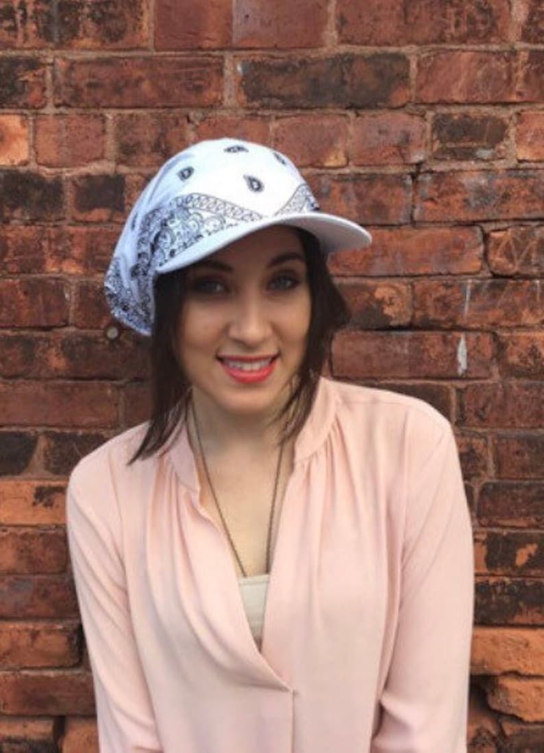 May include: A white bandana style baseball cap with a black paisley pattern. The cap is worn by a person with long brown hair and they are wearing a light pink shirt.