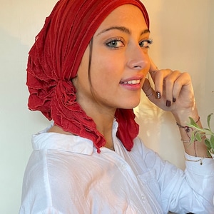 May include: A woman wearing a red ruffled head scarf and a white button-down shirt. She is looking to the side and smiling.