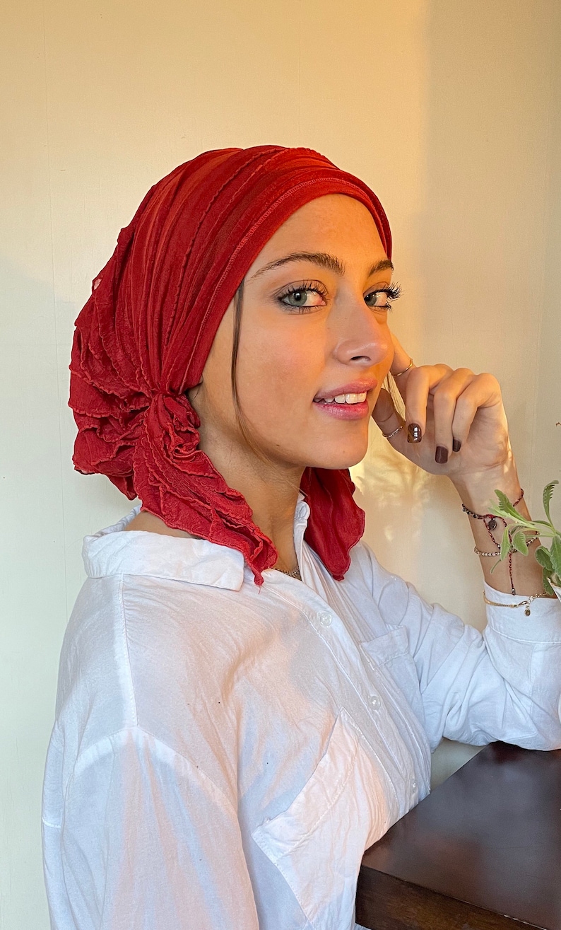 May include: A woman wearing a red ruffled head scarf and a white button-down shirt. She is looking to the side and smiling.