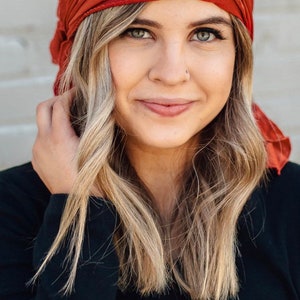 May include: A woman with long blonde hair is wearing a rust-colored headscarf. She is wearing a black long-sleeved shirt.