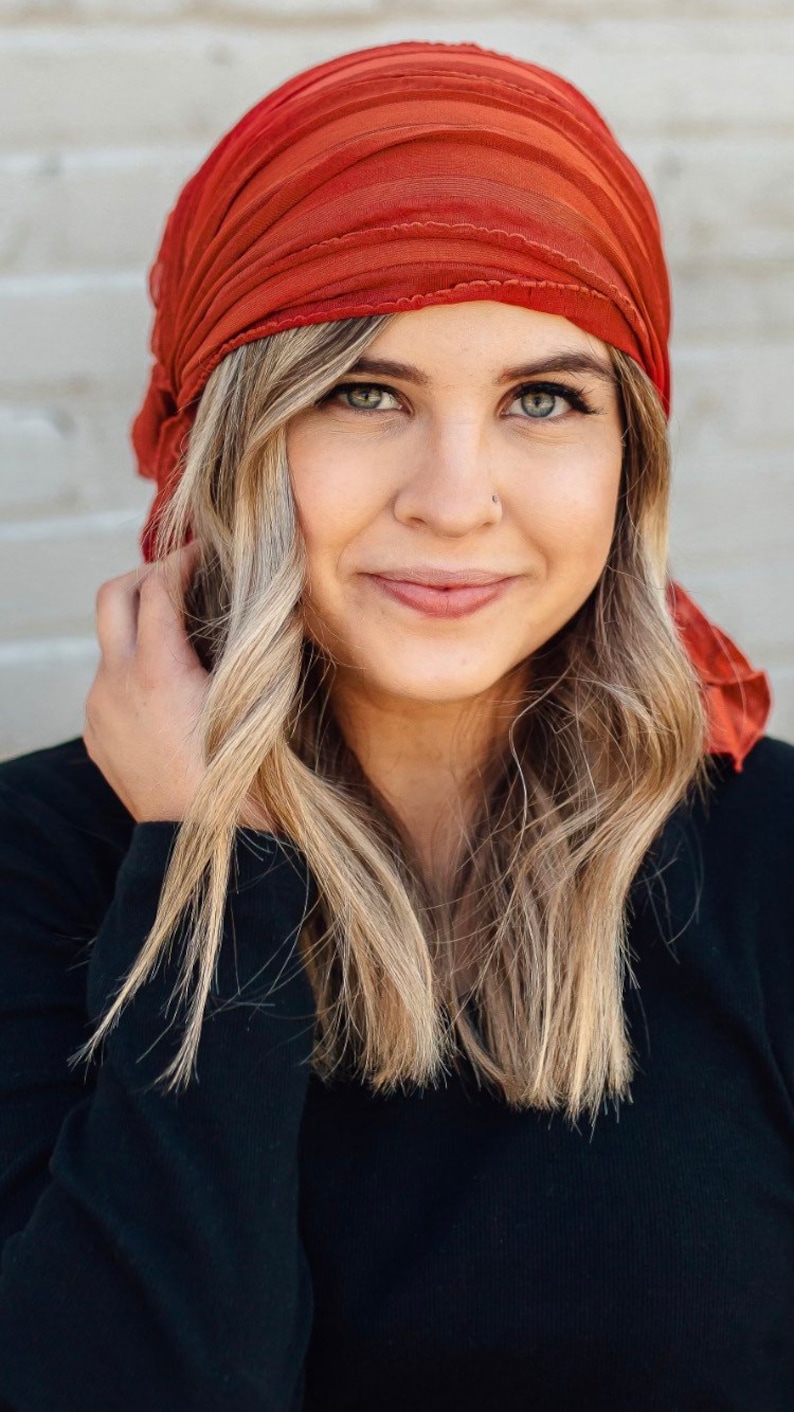 May include: A woman with long blonde hair is wearing a rust-colored headscarf. She is wearing a black long-sleeved shirt.