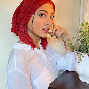 May include: A woman wearing a red ruffled turban and a white button-down shirt. She is leaning against a wooden table.