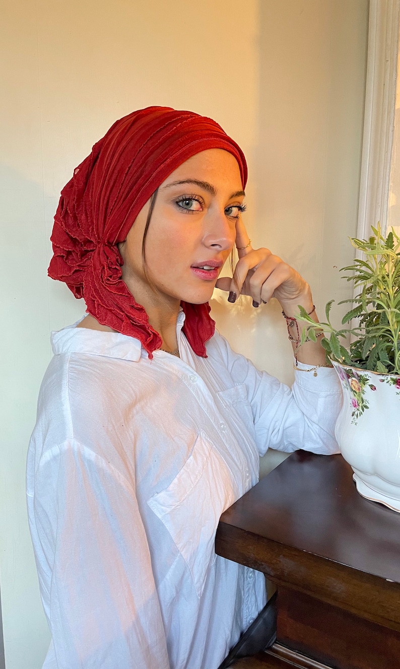May include: A woman wearing a red ruffled turban and a white button-down shirt. She is leaning against a wooden table.
