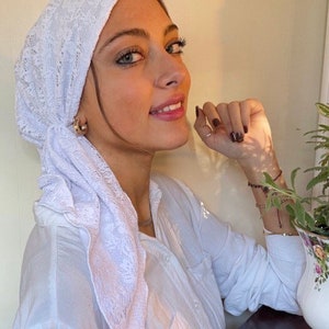 May include: A white lace headscarf with a delicate pattern. The scarf is tied in a knot at the back of the head and the ends are draped down the shoulders.