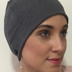 Puede incluir: Un gorro de lana gris oscuro hecho de tela suave. El gorro está puesto por una persona con una expresión neutral. El gorro tiene un diseño simple y es un accesorio versátil.