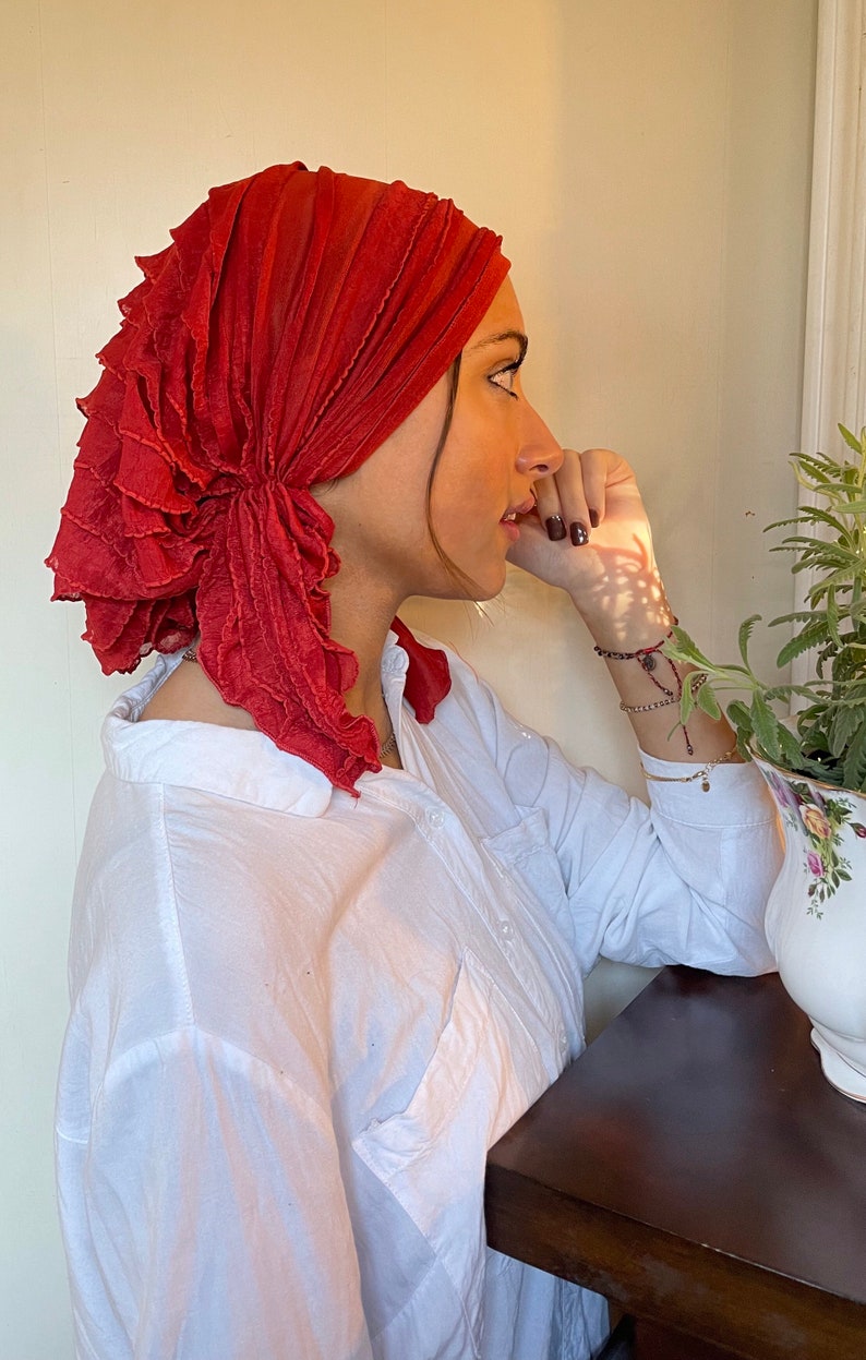 May include: A woman wearing a red ruffled turban. The turban is tied in a knot at the back of her head. She is wearing a white button-down shirt and has her hand on her chin.