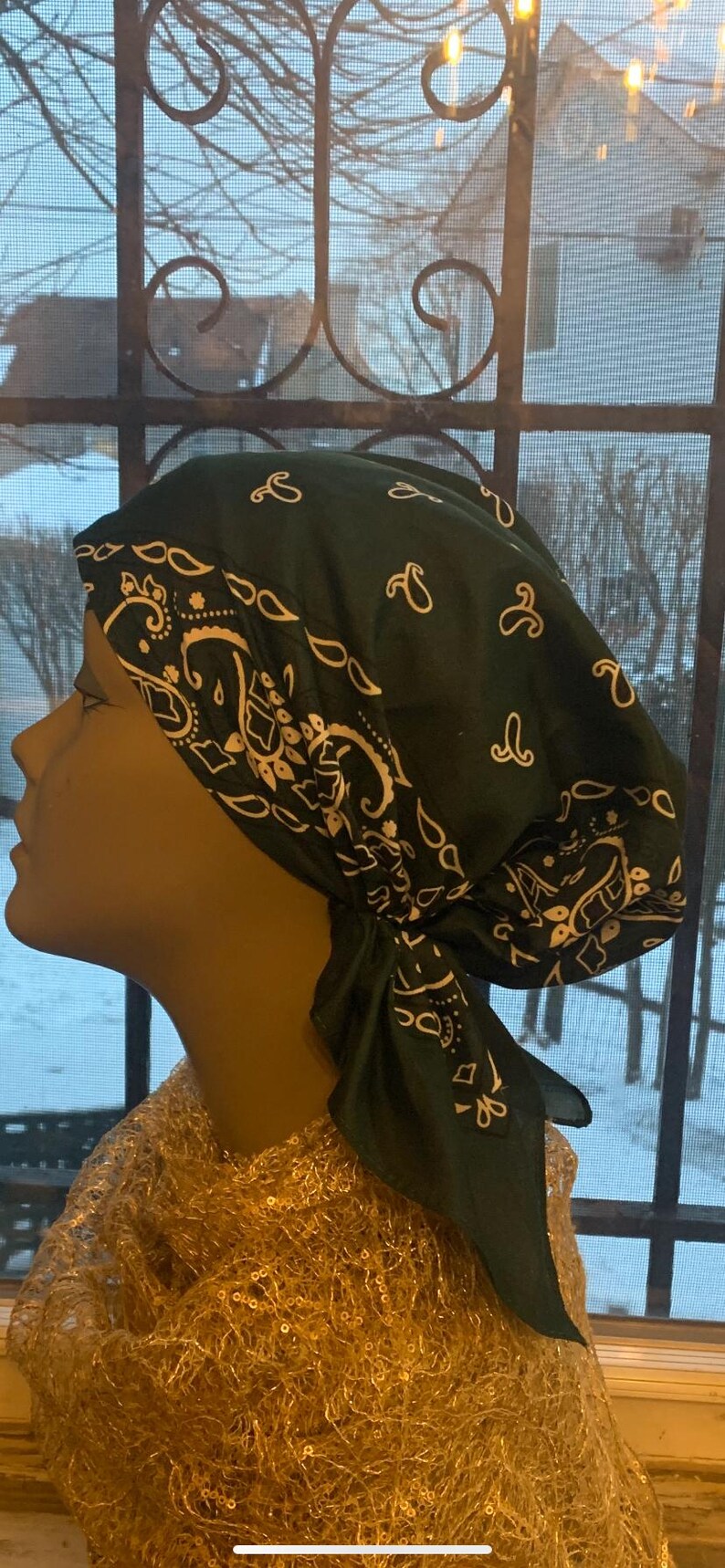 May include: A dark green bandana-style head covering with a white paisley pattern. The head covering is tied at the back and is displayed on a mannequin head. The mannequin is wearing a gold sequined top.
