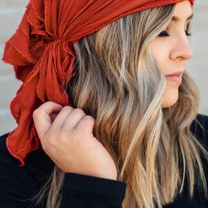 May include: A rust-colored fabric head scarf with a ruffled edge is tied around a person's head. The scarf is folded and tied in a way that creates a loose, flowing look.