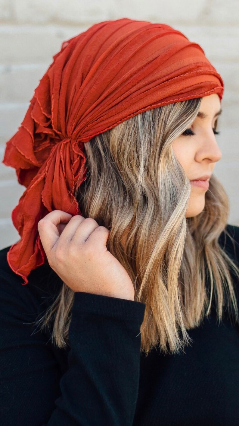 May include: A rust-colored fabric head scarf with a ruffled edge is tied around a person's head. The scarf is folded and tied in a way that creates a loose, flowing look.