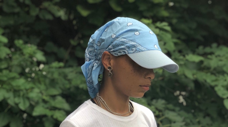 May include: A light blue bandana tied around a person's head with a light blue baseball cap on top. The bandana has a white polka dot pattern and a paisley design.