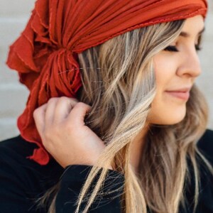 May include: A woman with long blonde hair is wearing a rust-colored headscarf tied in a knot at the back of her head. She is wearing a black long-sleeved shirt.