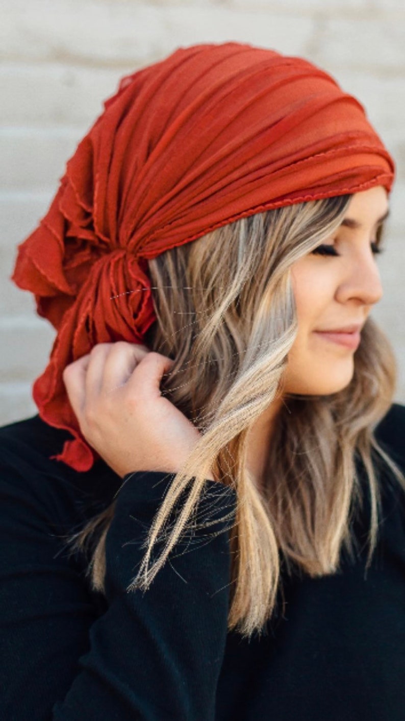 May include: A woman with long blonde hair is wearing a rust-colored headscarf tied in a knot at the back of her head. She is wearing a black long-sleeved shirt.