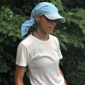 May include: A person wearing a light blue bandana tied around their head as a headband. The bandana has a white pattern. The person is also wearing a light blue baseball cap with a white brim. The person is wearing a white t-shirt.