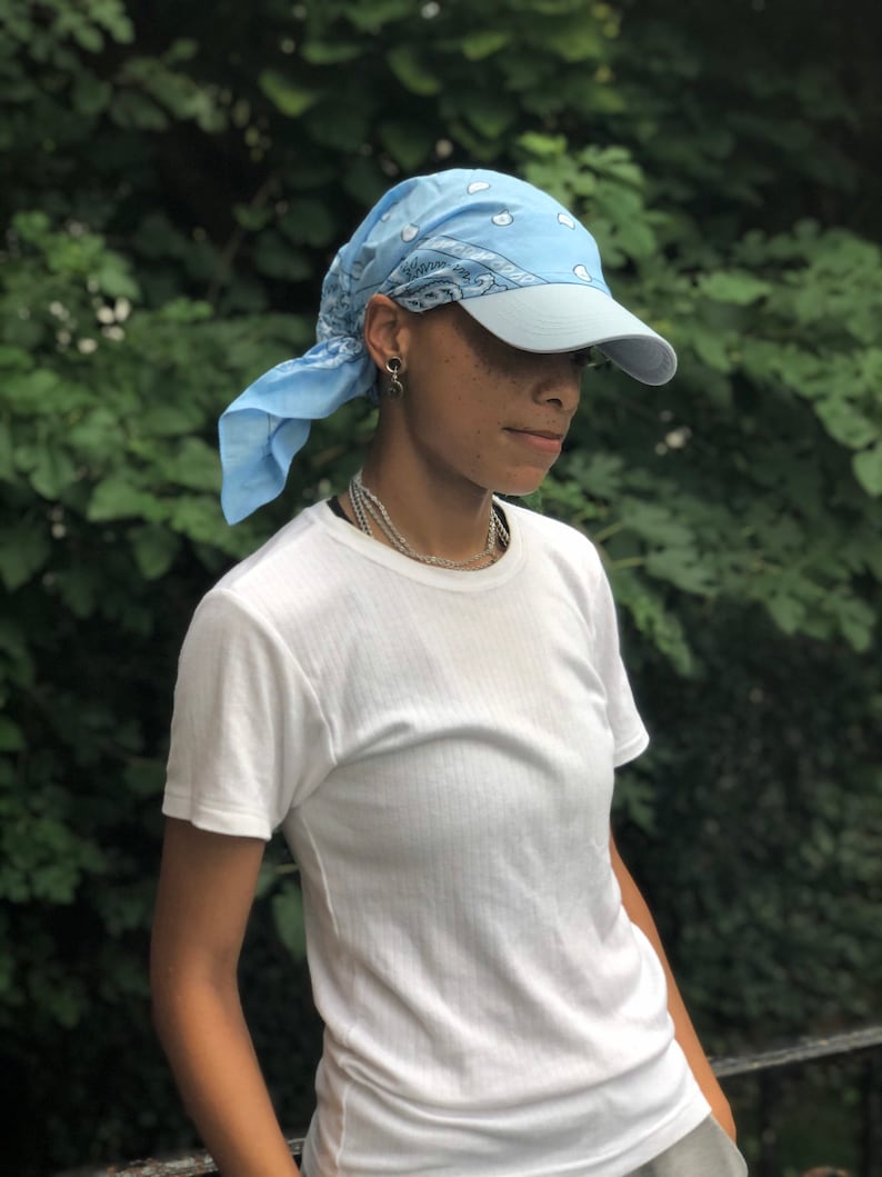 May include: A person wearing a light blue bandana tied around their head as a headband. The bandana has a white pattern. The person is also wearing a light blue baseball cap with a white brim. The person is wearing a white t-shirt.