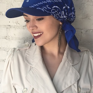 May include: A blue bandana with a white paisley pattern is tied around the head and worn as a cap. The bandana is worn under a blue baseball cap with a curved brim.