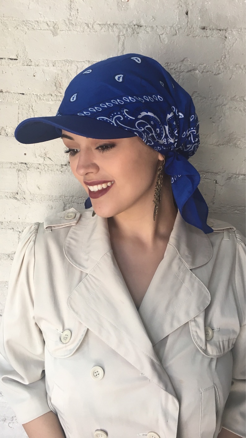 May include: A blue bandana with a white paisley pattern is tied around the head and worn as a cap. The bandana is worn under a blue baseball cap with a curved brim.