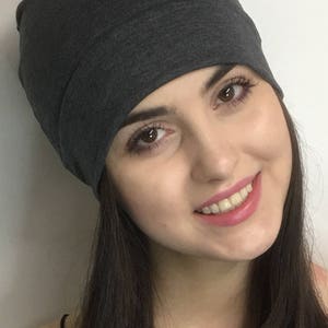 May include: A dark gray beanie hat made of soft fabric. The hat is worn on the head. The person wearing the hat has dark hair and is smiling. The background is a plain white wall.