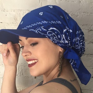 May include: A blue bandana with a white paisley pattern is tied around a person's head and tied into a knot at the back of the neck. The bandana is worn under a blue baseball cap with a visor.