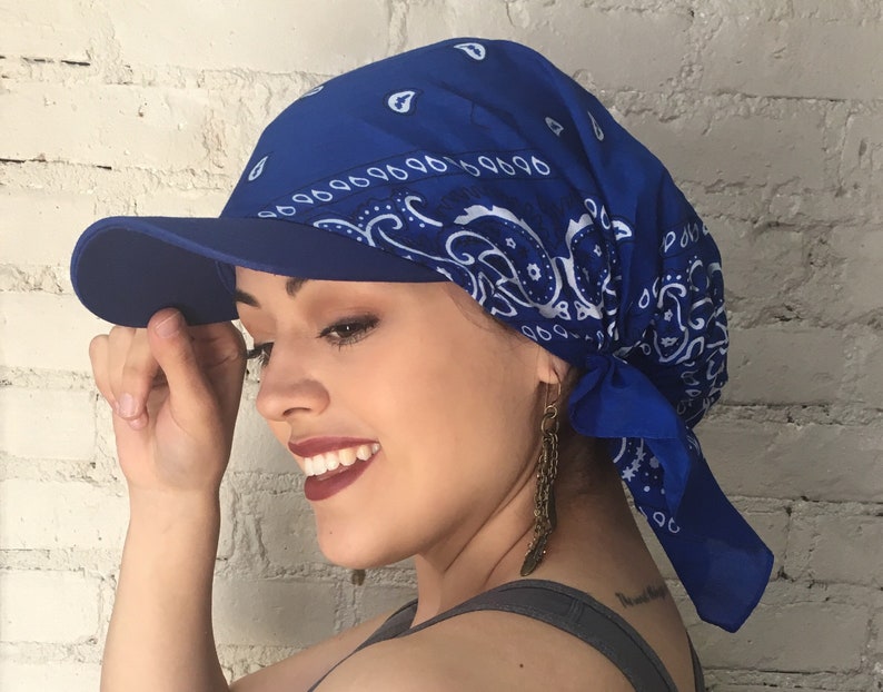May include: A blue bandana with a white paisley pattern is tied around a person's head and tied into a knot at the back of the neck. The bandana is worn under a blue baseball cap with a visor.