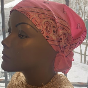 May include: A pink paisley bandana tied on a mannequin head. The bandana is a vibrant pink with a repeating paisley pattern in a lighter pink. A gold-colored, textured scarf is draped around the mannequin's neck.