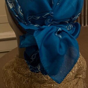 May include: A blue bandana head covering with a white paisley pattern, tied at the back. The head covering is draped over a mannequin head, with a gold-colored decorative element at the base.
