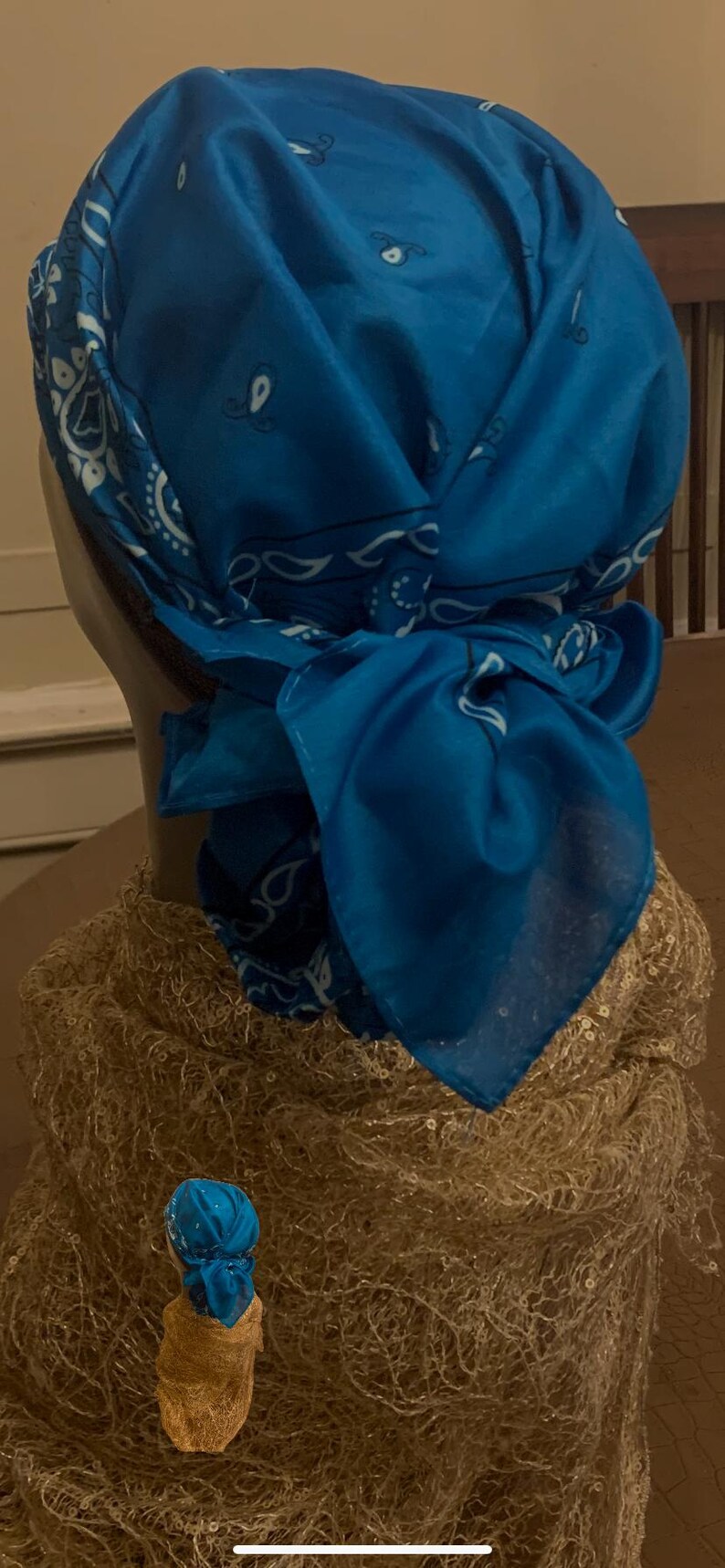 May include: A blue bandana head covering with a white paisley pattern, tied at the back. The head covering is draped over a mannequin head, with a gold-colored decorative element at the base.
