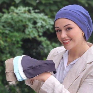 Puede incluir: Una mujer con una boina azul sostiene una pila de cuatro boinas de diferentes colores. Las boinas son en tonos de marrón, azul claro, blanco y negro.