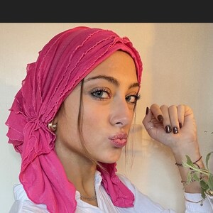 May include: A pink ruffled turban with a flower detail. The turban is worn by a person with dark hair and brown eyes. The person is wearing a white shirt.