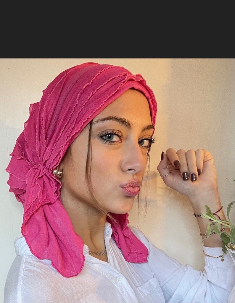 May include: A pink ruffled turban with a flower detail. The turban is worn by a person with dark hair and brown eyes. The person is wearing a white shirt.