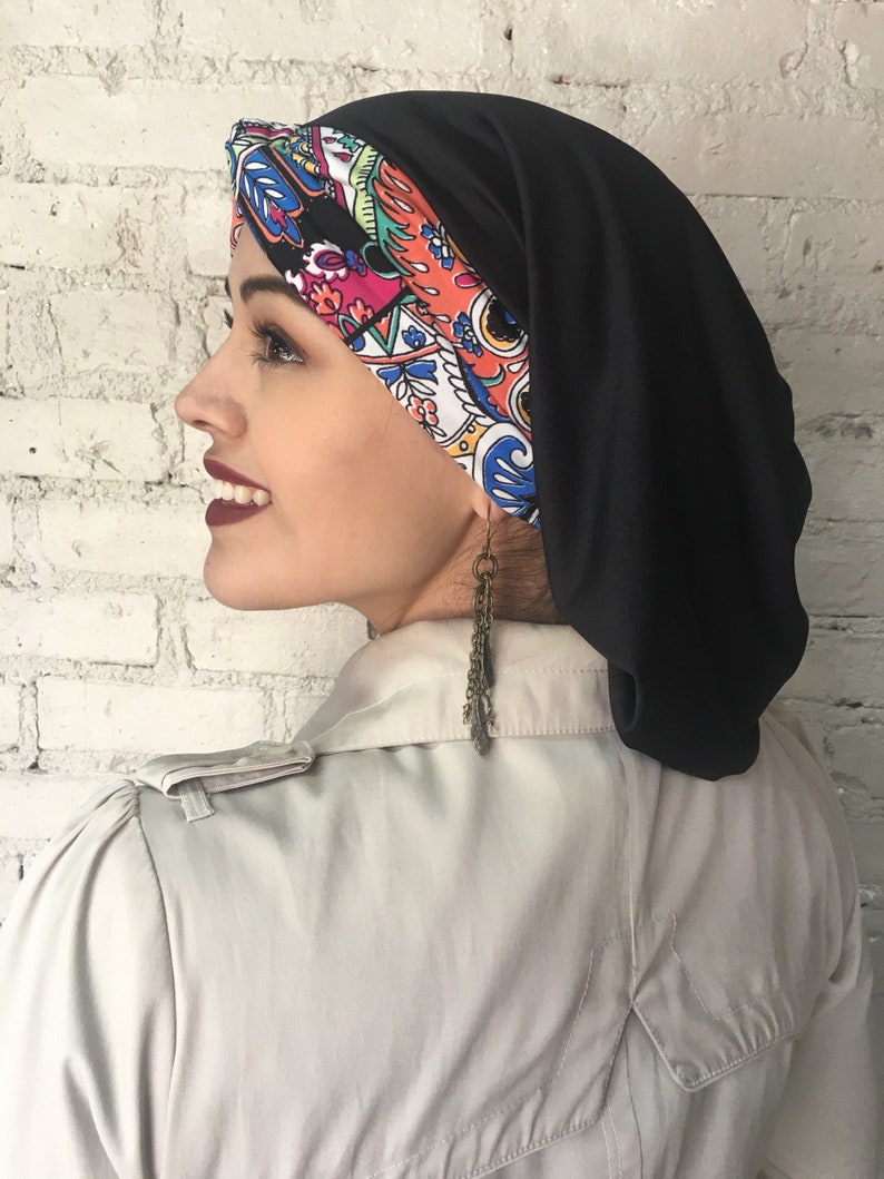 K&ouml;nnte beinhalten: Ein schwarzer und farbenfroher Turban-Kopfbedeckung mit Muster. Die Kopfbedeckung besteht aus einem weichen, dehnbaren Stoff. Der Stoff ist au&szlig;en schwarz und innen mit einem farbenfrohen Paisley-Muster versehen.