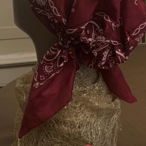 May include: A burgundy bandana head covering with a white paisley pattern and decorative swirls. The bandana is tied at the nape of the neck. A smaller version of the bandana is also present.
