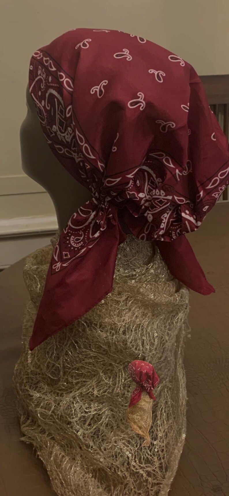 May include: A burgundy bandana head covering with a white paisley pattern and decorative swirls. The bandana is tied at the nape of the neck. A smaller version of the bandana is also present.
