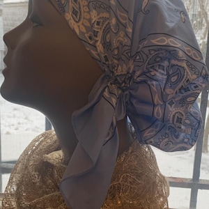 May include: A light blue paisley print headscarf tied at the back, displayed on a mannequin head. The headscarf is made of a soft fabric and features a classic paisley design in white and gray. A gold-colored, textured scarf is draped around the mannequin's neck.