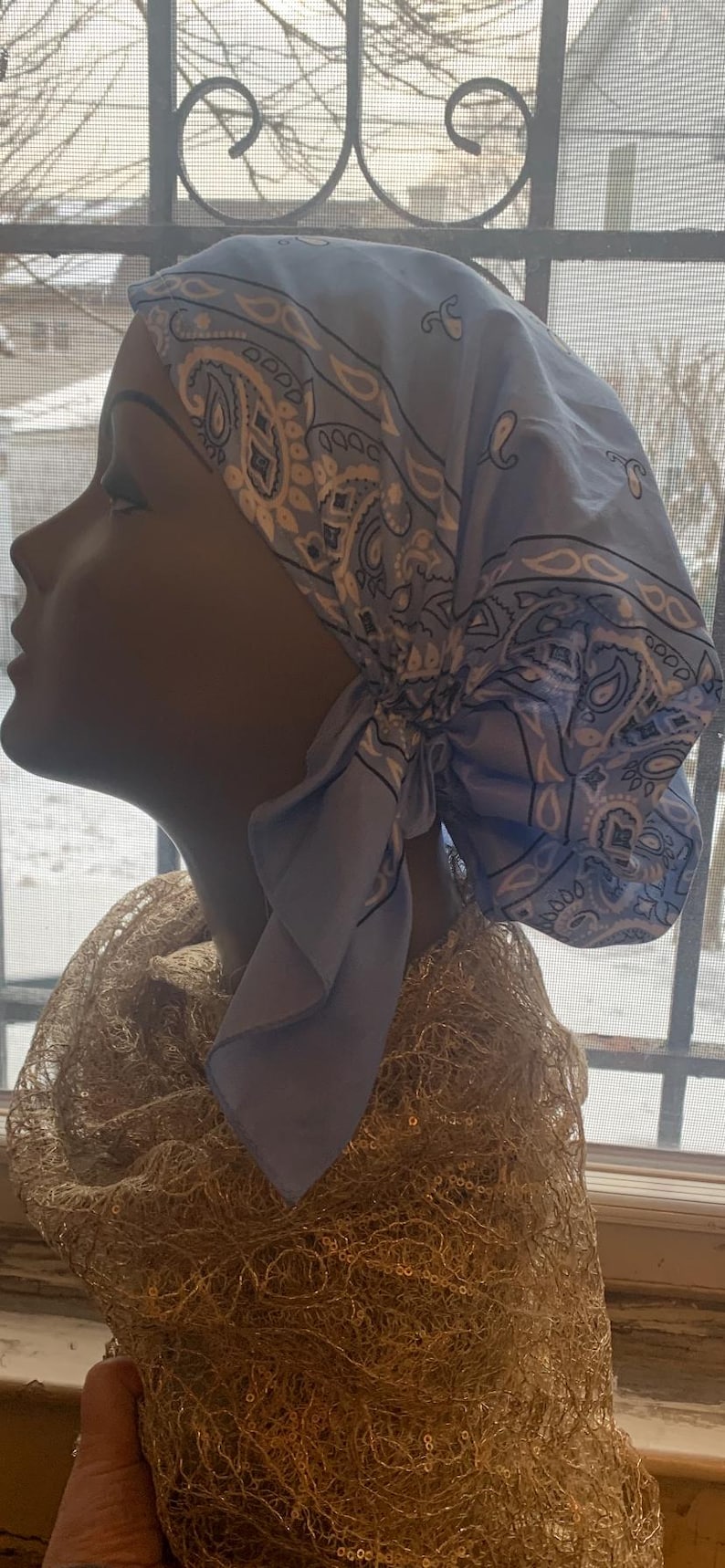 May include: A light blue paisley print headscarf tied at the back, displayed on a mannequin head. The headscarf is made of a soft fabric and features a classic paisley design in white and gray. A gold-colored, textured scarf is draped around the mannequin's neck.